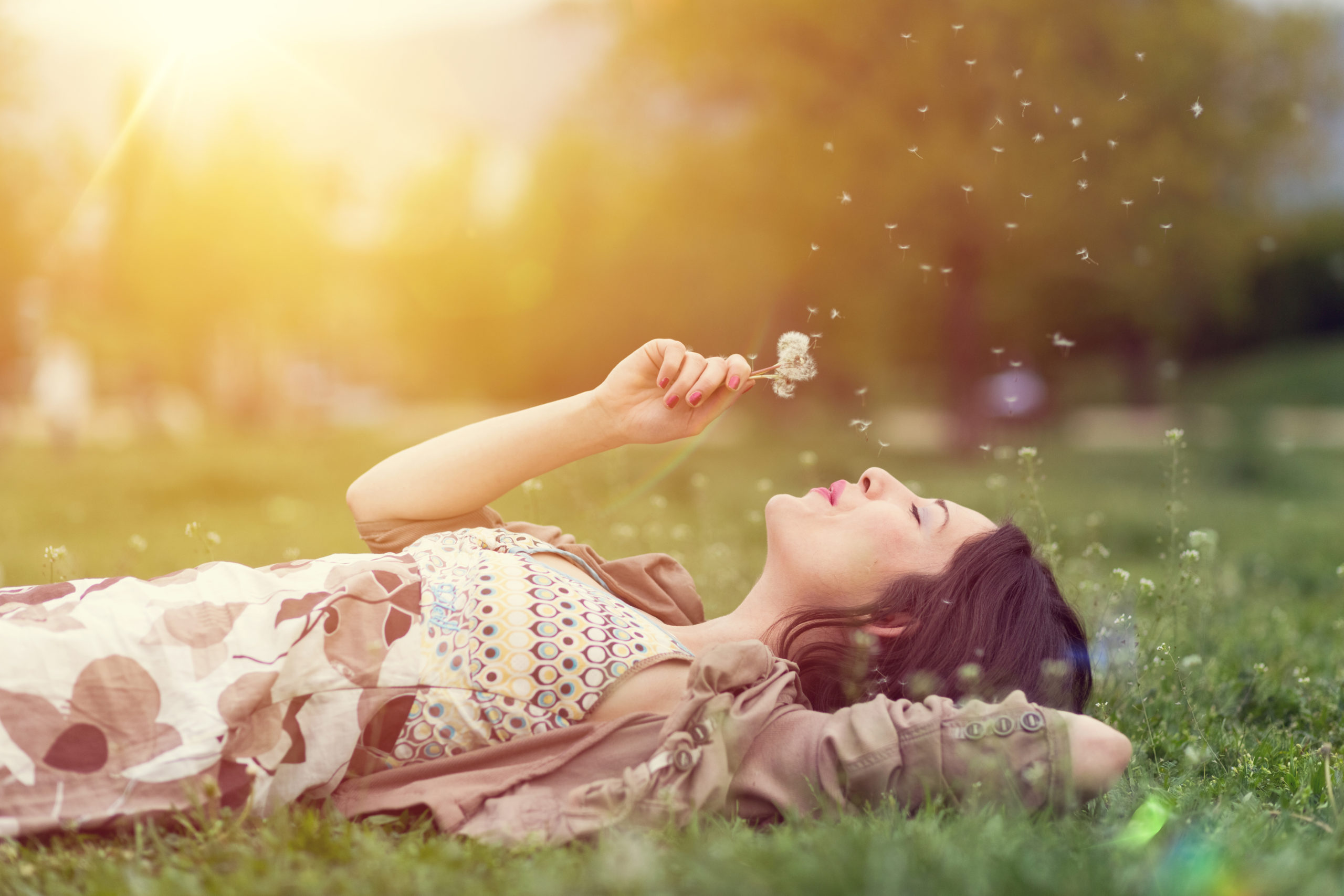 Image shows woman lying on the grass blowing on a dandelion