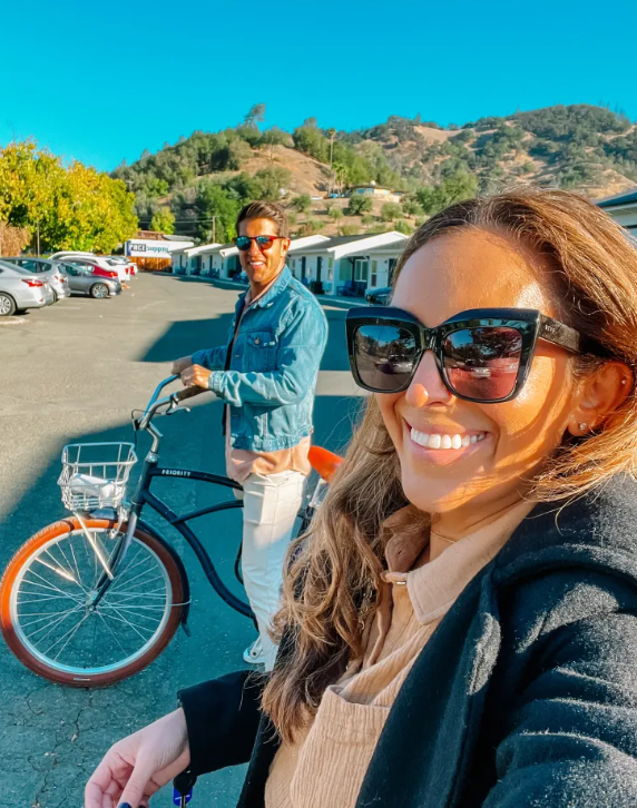image shows a man riding a bike and a woman taking a selfie of the two of them with the hotel in the background